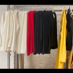 MIRANEKS - Rząd ubrań w różnych kolorach (biały, czerwony, czarny, żółty) wiszących na wieszakach na tle białej ceglanej ściany.