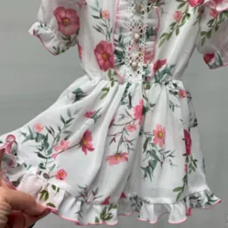 Biała, zwiewna sukienka dziewczęca w r&oacute;żowe kwiaty, z falbankami i koronkowym wykończeniem, wisząca na wieszaku i przytrzymywana przez dłoń.