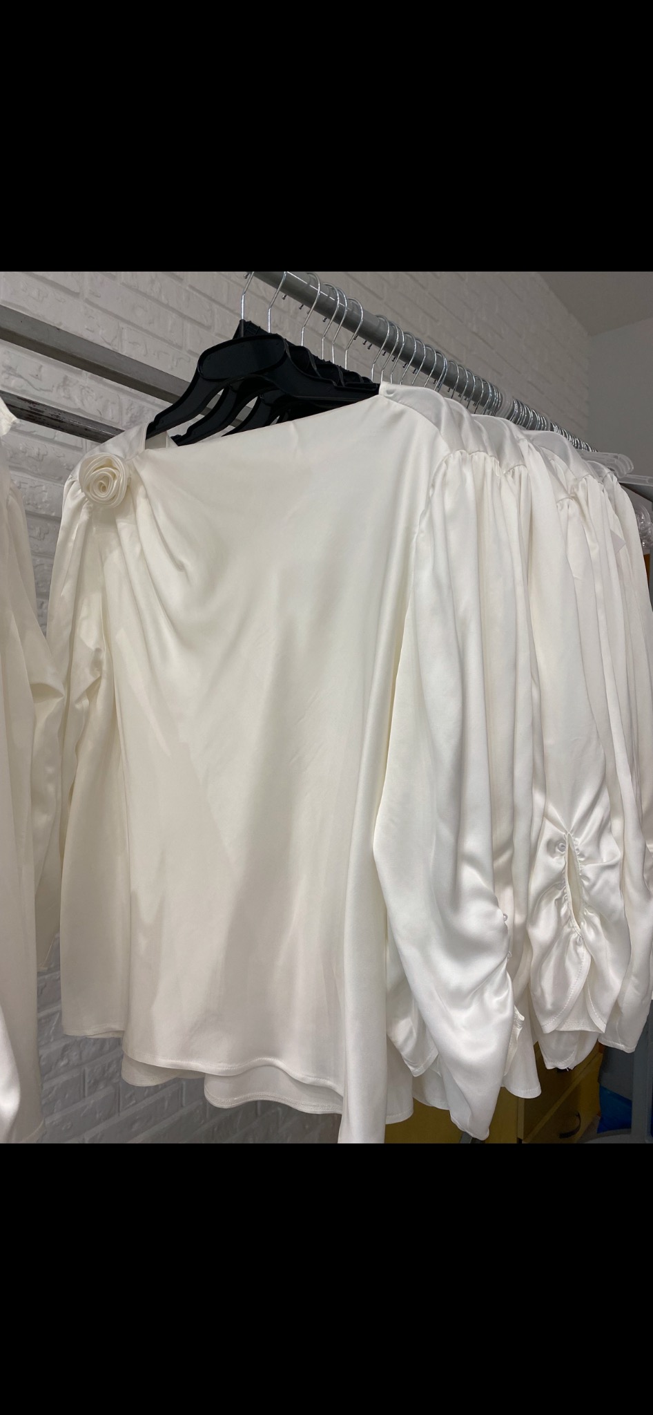 Rząd białych, satynowych bluzek damskich z ozdobnymi kwiatami na ramieniu, wiszących na czarnych wieszakach w szwalni w Łasku, na tle ściany z białej cegły.