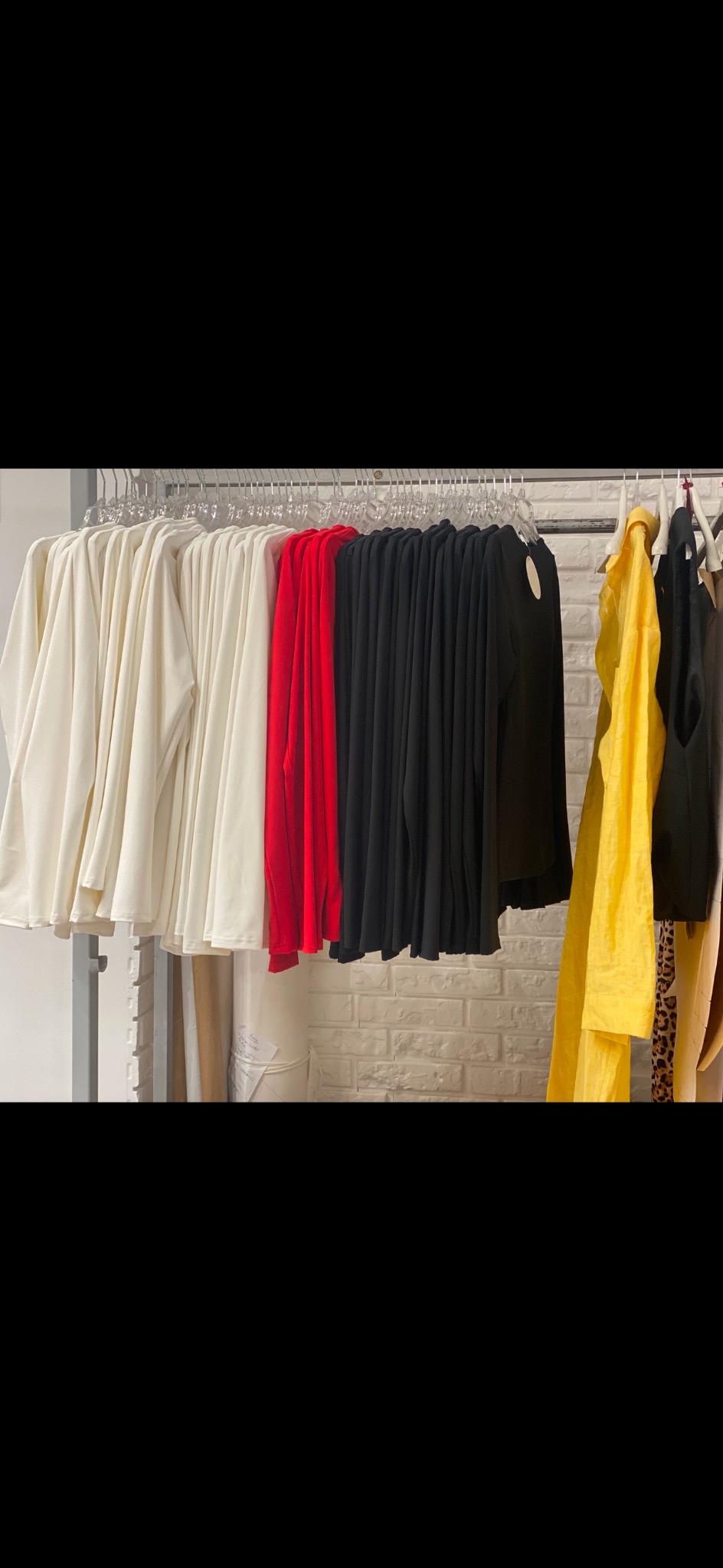 Rząd ubrań w różnych kolorach (biały, czerwony, czarny, żółty) wiszących na wieszakach na tle białej ceglanej ściany.