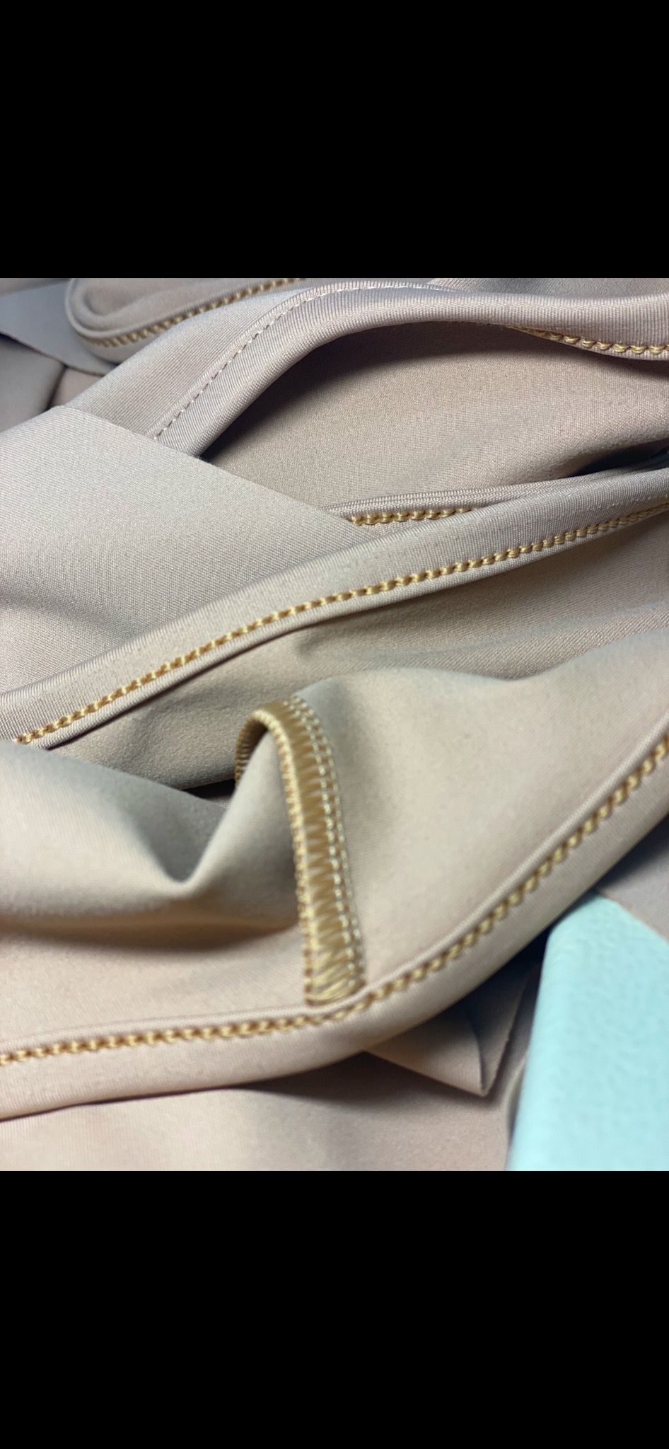 Beżowa tkanina z ozdobnym, złotym szwem maszynowym, widoczny fragment jasnoniebieskiego materiału w rogu kadru.