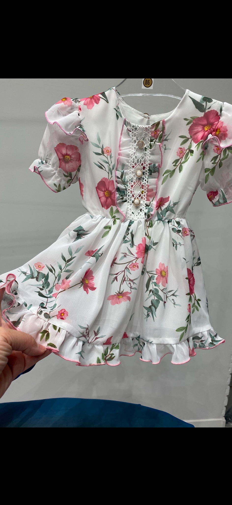 Biała, zwiewna sukienka dziewczęca w różowe kwiaty, z falbankami i koronkowym wykończeniem, wisząca na wieszaku i przytrzymywana przez dłoń.