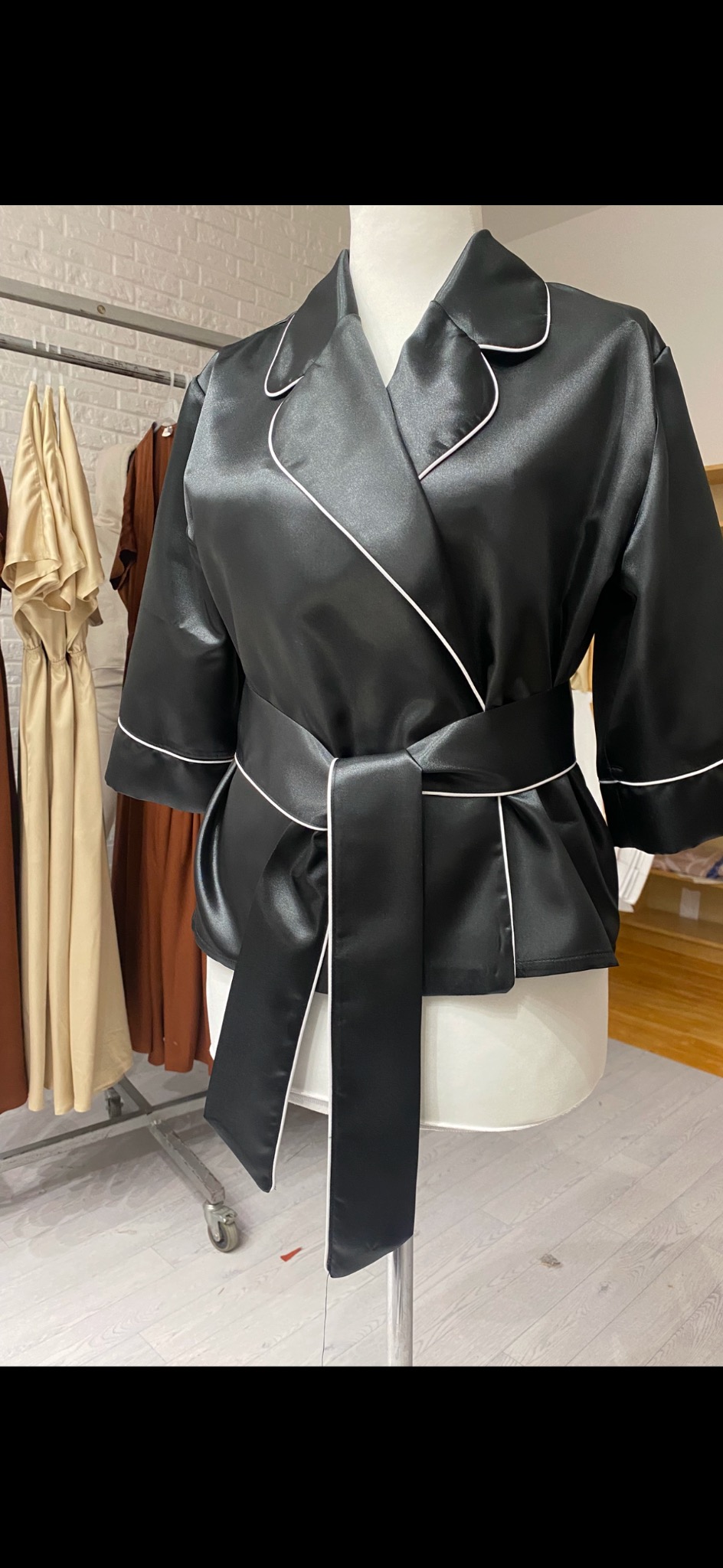 Czarna satynowa bluzka damska z białym obszyciem i wiązanym paskiem, prezentowana na manekinie na tle wieszaków z odzieżą.