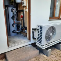 Zewnętrzna jednostka pompy ciepła marki Beretta zamontowana na betonowych bloczkach, z widocznym zbiornikiem akumulacyjnym w pomieszczeniu obok.