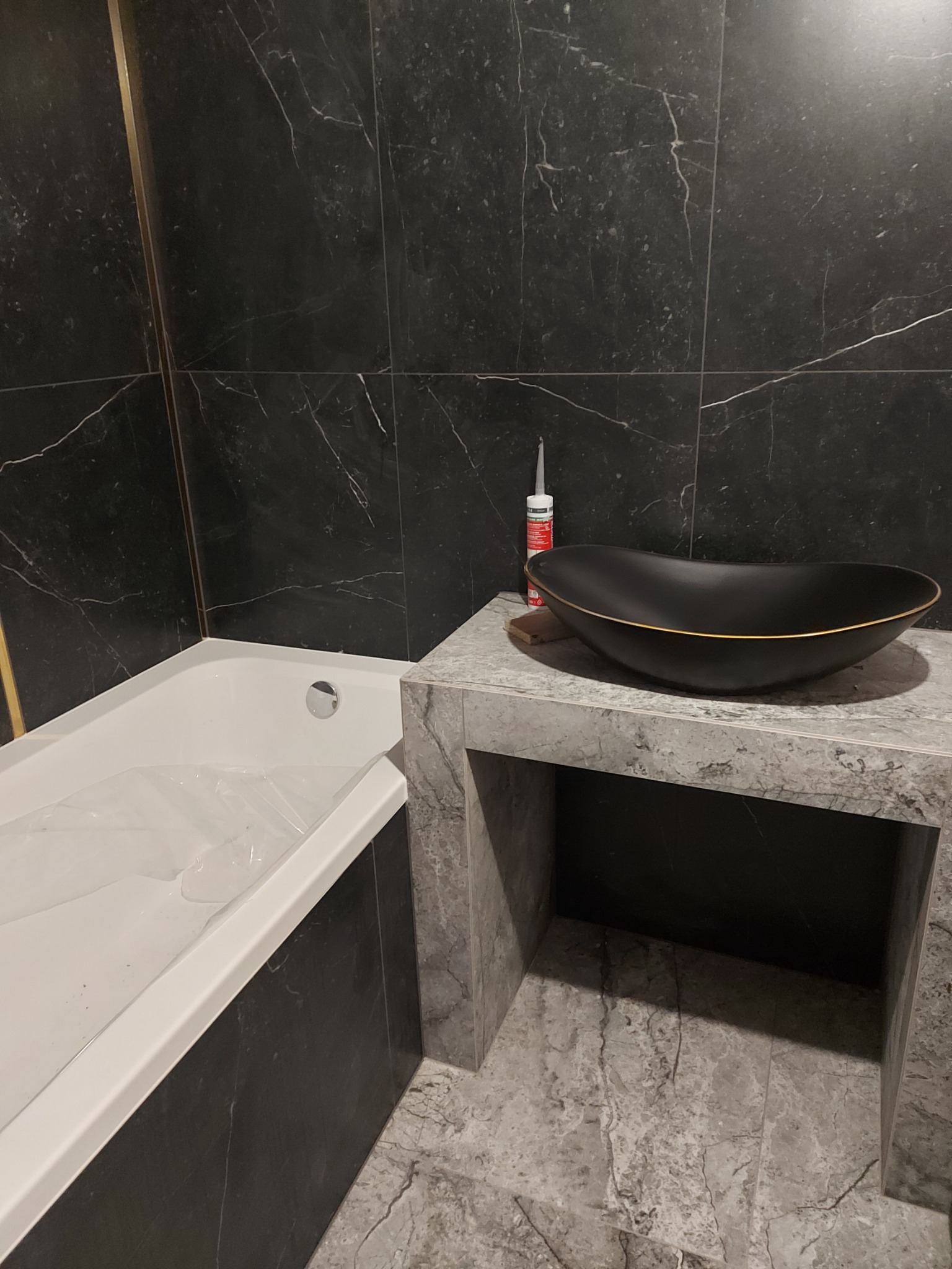 Czarna, owalna umywalka z delikatnym złotym obramowaniem na blacie z szarego marmuru, obok biała wanna z czarnymi panelami i czarne marmurowe płytki ścienne z delikatnymi złotymi akcentami...