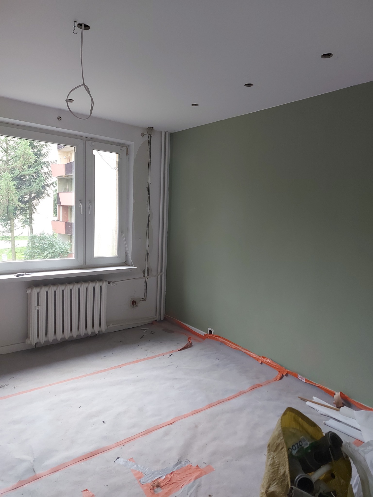 Pokój w trakcie prac malarskich: ściana pomalowana na oliwkowy kolor, zabezpieczona podłoga folią, widoczne przewody elektryczne i grzejnik.