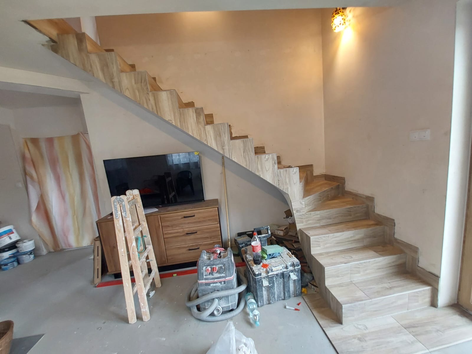 Wykończone schody z płytek imitujących drewno w trakcie prac wykończeniowych; widoczne narzędzia i drabina, a w tle meble i telewizor.