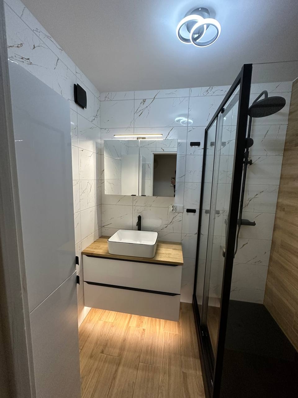 Nowoczesna łazienka z biało-czarnymi akcentami: kabina prysznicowa typu walk-in z czarną ramą, umywalka nablatowa na drewnianym blacie, białe płytki imitujące marmur na ścianach i drewnopodobna...