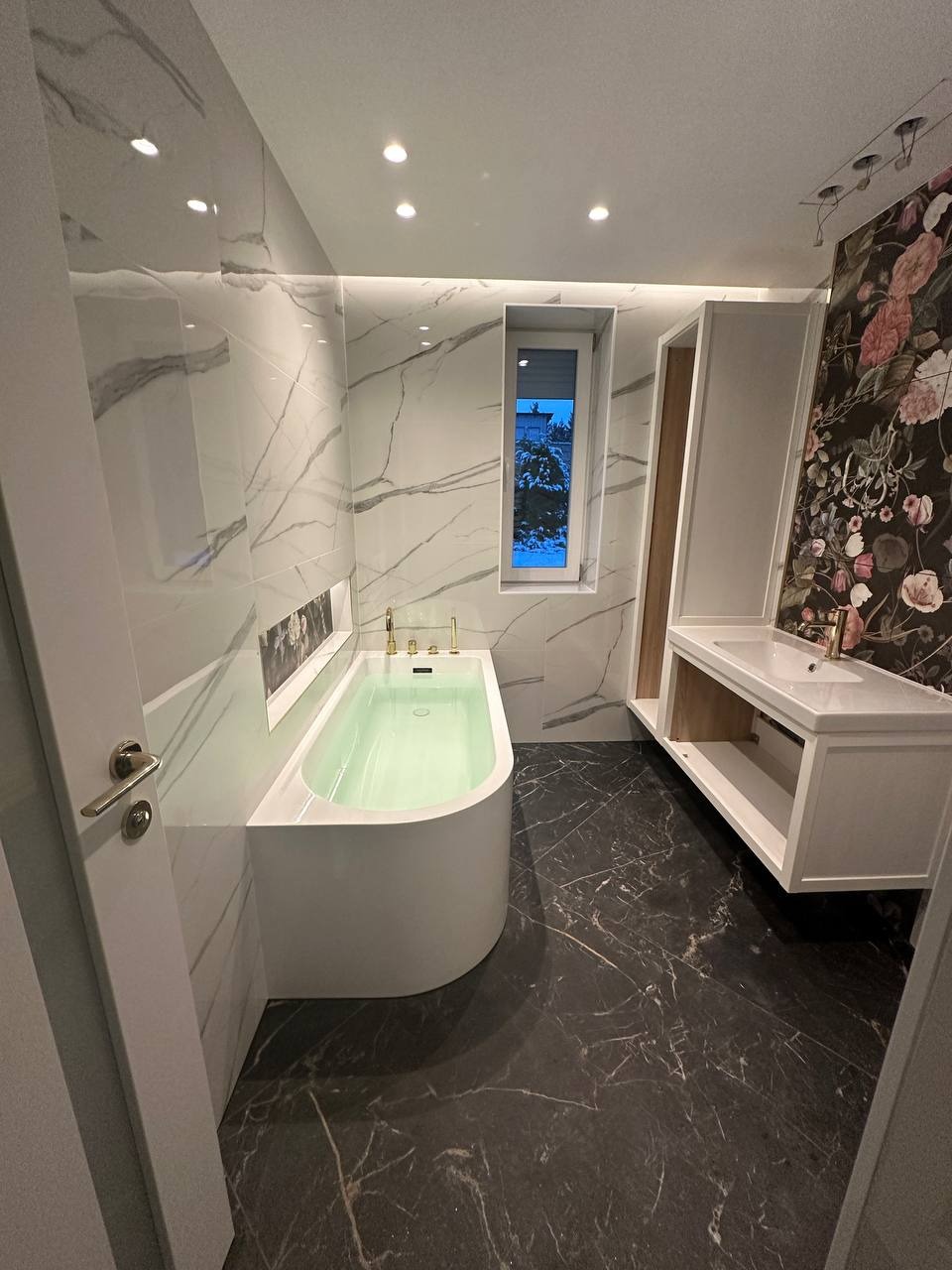 Nowoczesna łazienka z białą wanną wypełnioną wodą, marmurowe płytki na ścianach, ciemna podłoga z żyłkowaniem, biała szafka pod umywalką i ściana z motywem kwiatowym.