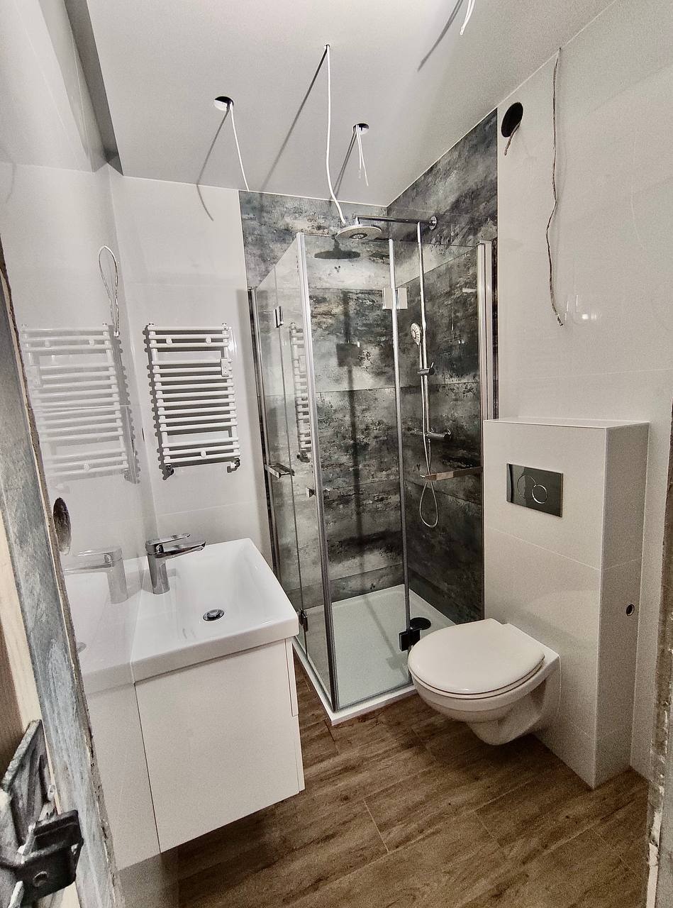 Nowoczesna łazienka w trakcie remontu: kabina prysznicowa z szarymi płytkami imitującymi kamień, biała umywalka z szafką, podwieszana toaleta, grzejnik drabinkowy, widoczne kable wystające z sufitu.