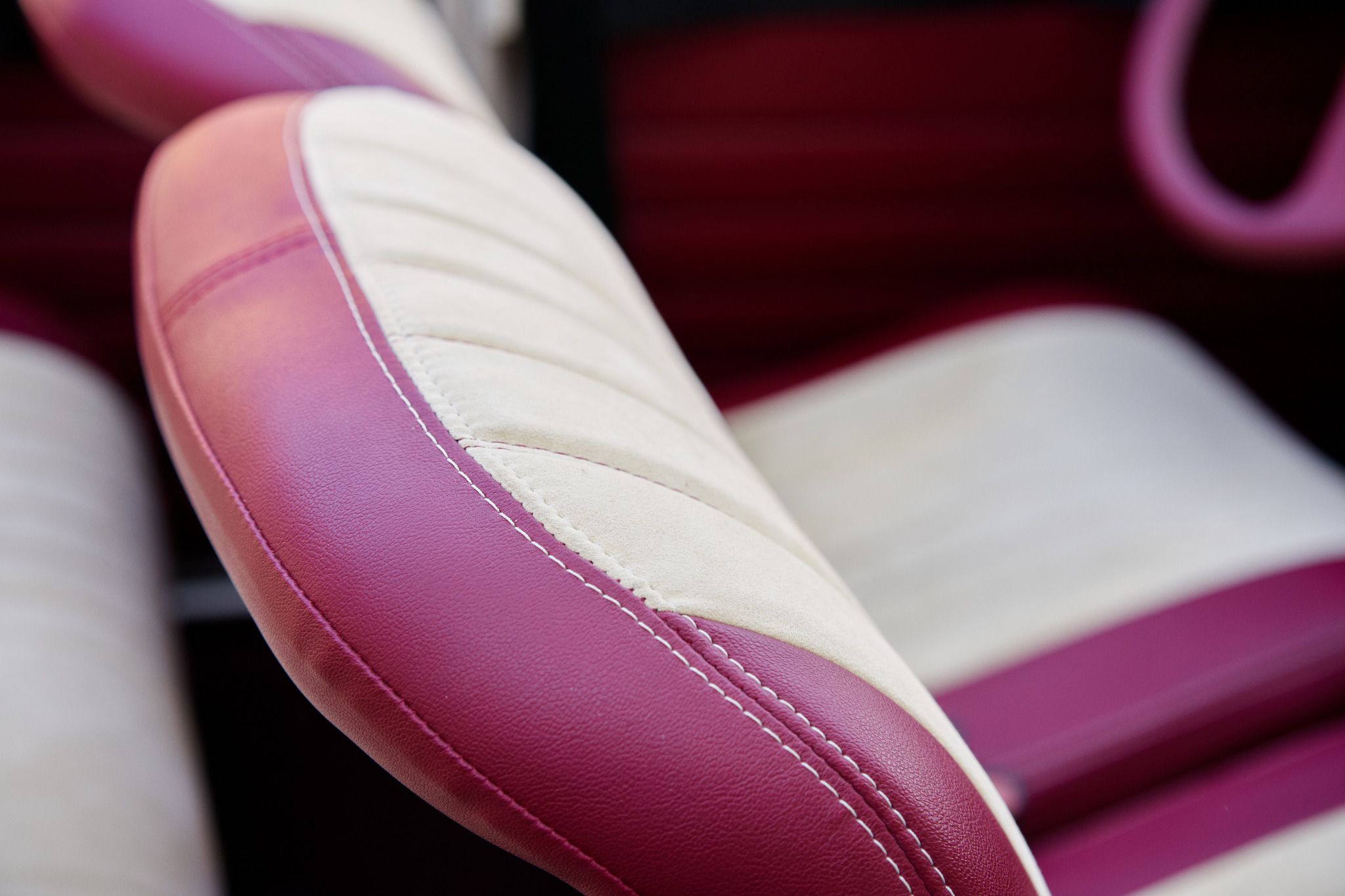 Fragment fotela samochodowego z bordową skórą i kremową alcantarą, widoczne precyzyjne szwy i fragment kierownicy w tle.