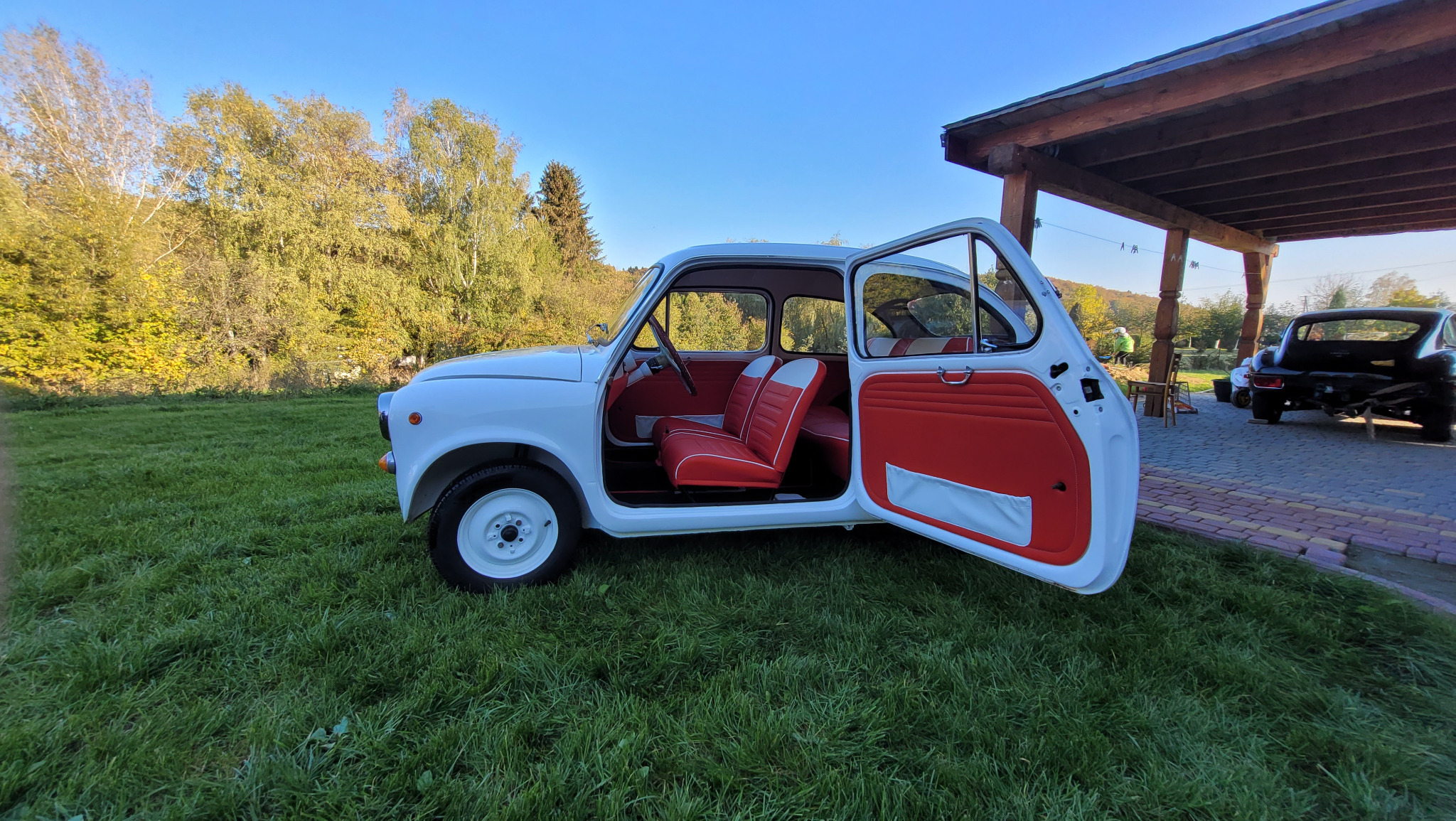 Biały, odrestaurowany samochód Zastava 750 z otwartymi drzwiami, prezentujący czerwono-białą tapicerkę siedzeń i drzwi, zaparkowany na trawniku przed drewnianą wiatą.
