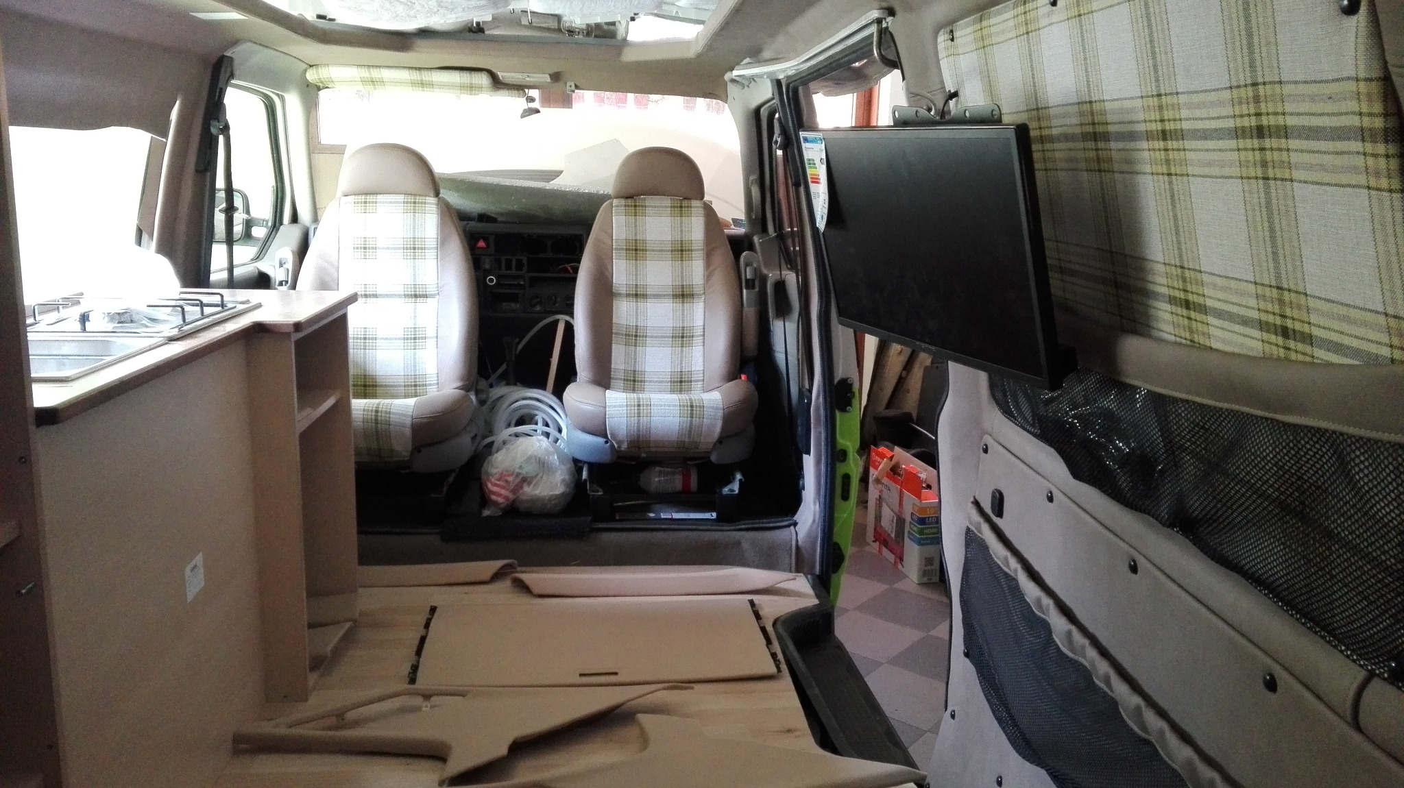 Wnętrze przerabianego busa na kampera: widoczne fotele z nową, kraciastą tapicerką, zabudowa kuchenna z kuchenką gazową, panel ścienny z zamontowanym telewizorem oraz elementy wykończeniowe podłogi...