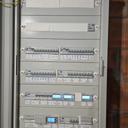 Bartosz Rokosz - Szara skrzynka rozdzielcza z bezpiecznikami, licznikami i wyłącznikami, zamontowana na ścianie z cegły. Widoczne oznaczenia obwodów i komponentów.