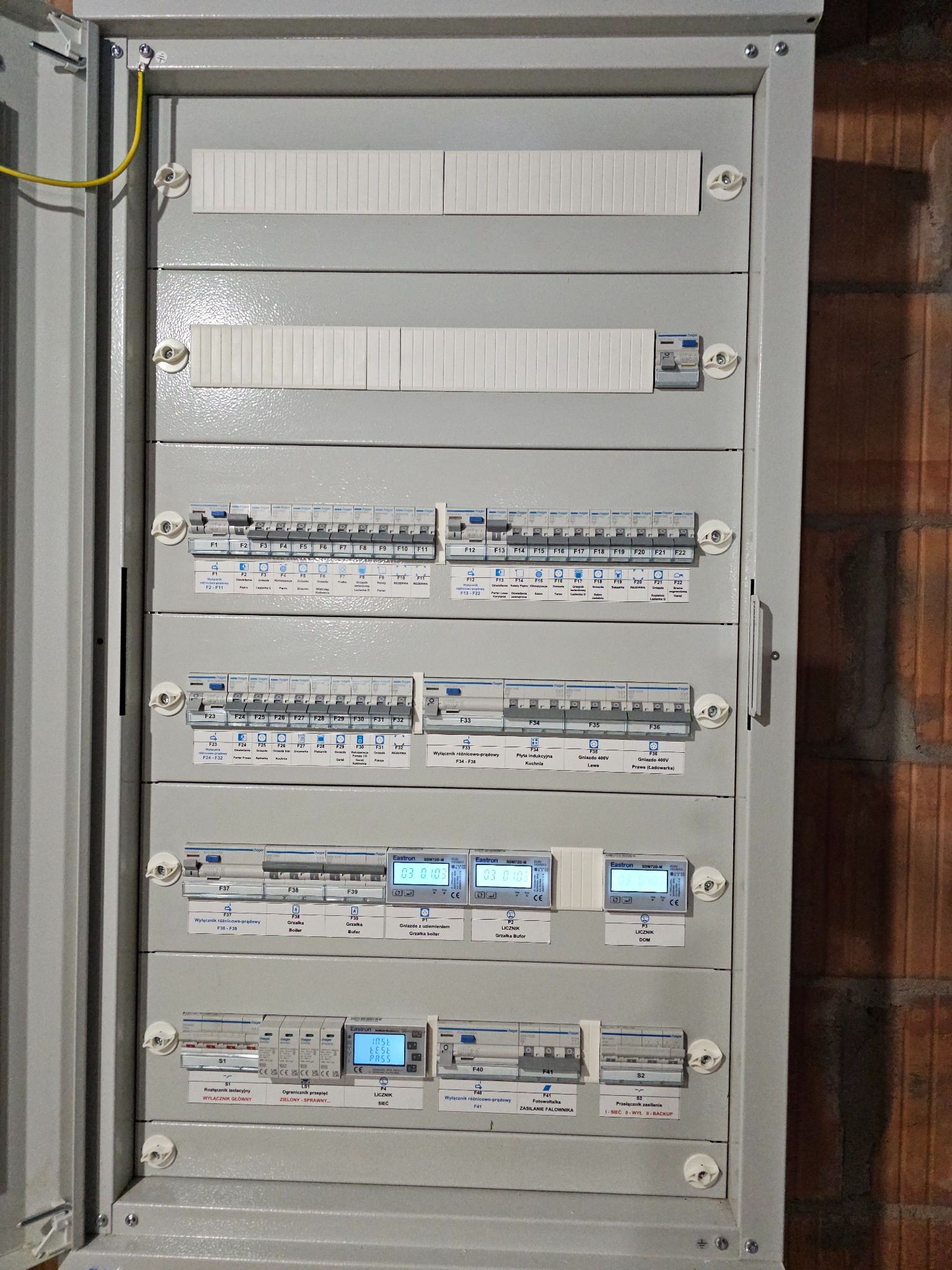 Szara skrzynka rozdzielcza z bezpiecznikami, licznikami i wyłącznikami, zamontowana na ścianie z cegły. Widoczne oznaczenia obwodów i komponentów.