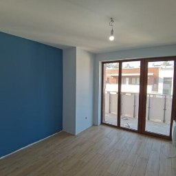 TOMI Remonty Wykończenia Wnętrz Brukarstwo - Pomalowane na niebiesko wnętrze pokoju z drewnopodobnymi panelami podłogowymi i balkonem z widokiem na inne budynki.