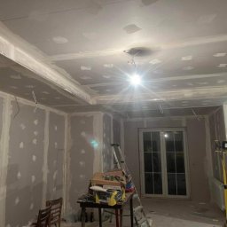 TOMI Remonty Wykończenia Wnętrz Brukarstwo - Pokój w trakcie wykańczania: ściany obłożone płytami kartonowo-gipsowymi z widocznymi łączeniami i szpachlowaniem, sufit z dekoracyjną zabudową z płyt gipsowych, okno balkonowe, drabina i narzędzia...