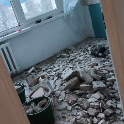 TOMI Remonty Wykończenia Wnętrz Brukarstwo - Pokój w trakcie skuwania posadzki, widoczne gruz, dwa zielone wiadra, wiadro z zaprawą, młotowiertarka i osłonięte folią okno.