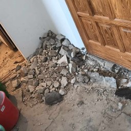 TOMI Remonty Wykończenia Wnętrz Brukarstwo - Fragment podłogi w trakcie skuwania starej posadzki, widoczne gruz, odłamki betonu, brud, wiadro i łom przy futrynie drzwi.