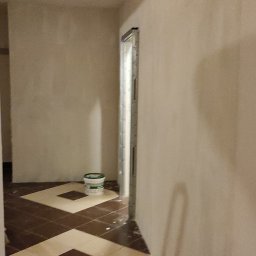 TOMI Remonty Wykończenia Wnętrz Brukarstwo - Przygotowane do malowania ściany z widoczną puszką farby w pomieszczeniu z ułożonymi płytkami podłogowymi w karo. W tle ościeżnica drzwiowa bez drzwi.