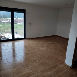 Jasne, puste pomieszczenie z ułożoną podłogą z paneli w odcieniach brązu, wzór jodełki, widoczne czarne przesuwne drzwi balkonowe, ściany przygotowane do malowania, widok na zewnątrz na trawnik...