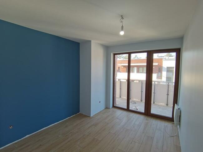 Pomalowane na niebiesko wnętrze pokoju z drewnopodobnymi panelami podłogowymi i balkonem z widokiem na inne budynki.