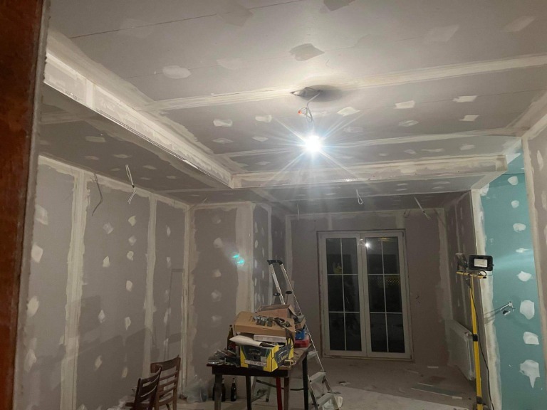Pokój w trakcie wykańczania: ściany obłożone płytami kartonowo-gipsowymi z widocznymi łączeniami i szpachlowaniem, sufit z dekoracyjną zabudową z płyt gipsowych, okno balkonowe, drabina i narzędzia...