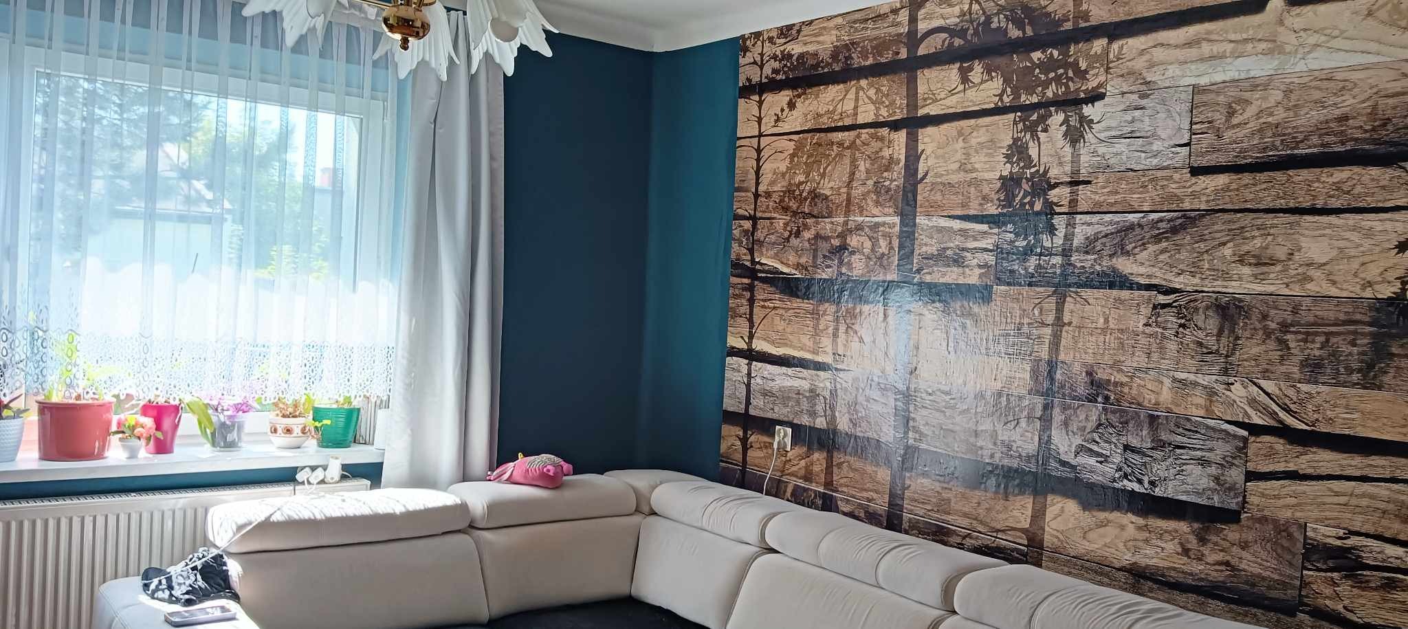 Pokój z ciemnymi ścianami, okno z firanką, biały narożnik i ściana z fototapetą imitującą drewniane deski z motywem drzew.