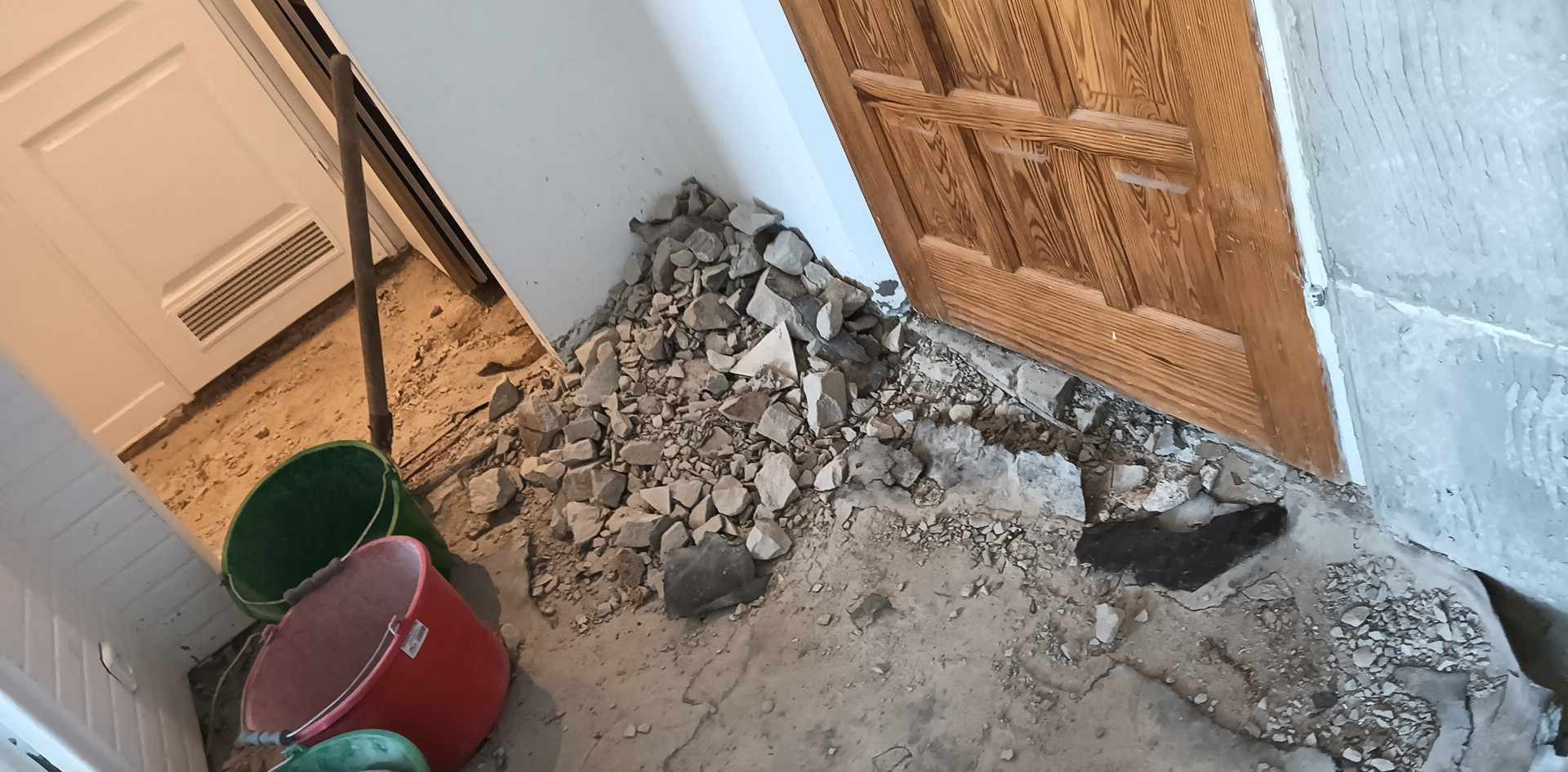 Fragment podłogi w trakcie skuwania starej posadzki, widoczne gruz, odłamki betonu, brud, wiadro i łom przy futrynie drzwi.