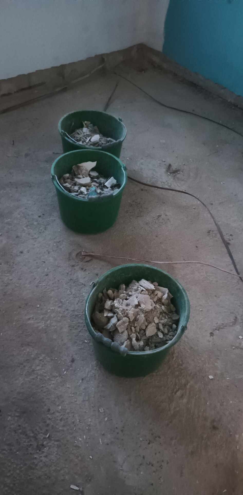 Trzy zielone wiadra wypełnione gruzem i resztkami materiałów budowlanych stoją na szarej, betonowej podłodze w trakcie prac remontowych.