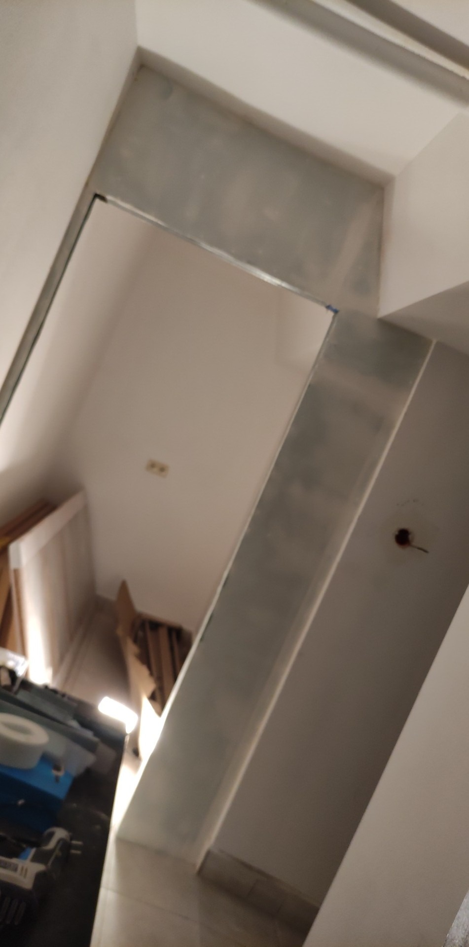 Skośne ujęcie surowej ściany z nałożoną szarą warstwą, prawdopodobnie gładzi, widoczne ślady po szpachlowaniu i niezabezpieczone gniazdko elektryczne, w tle narzędzia i materiały budowlane.