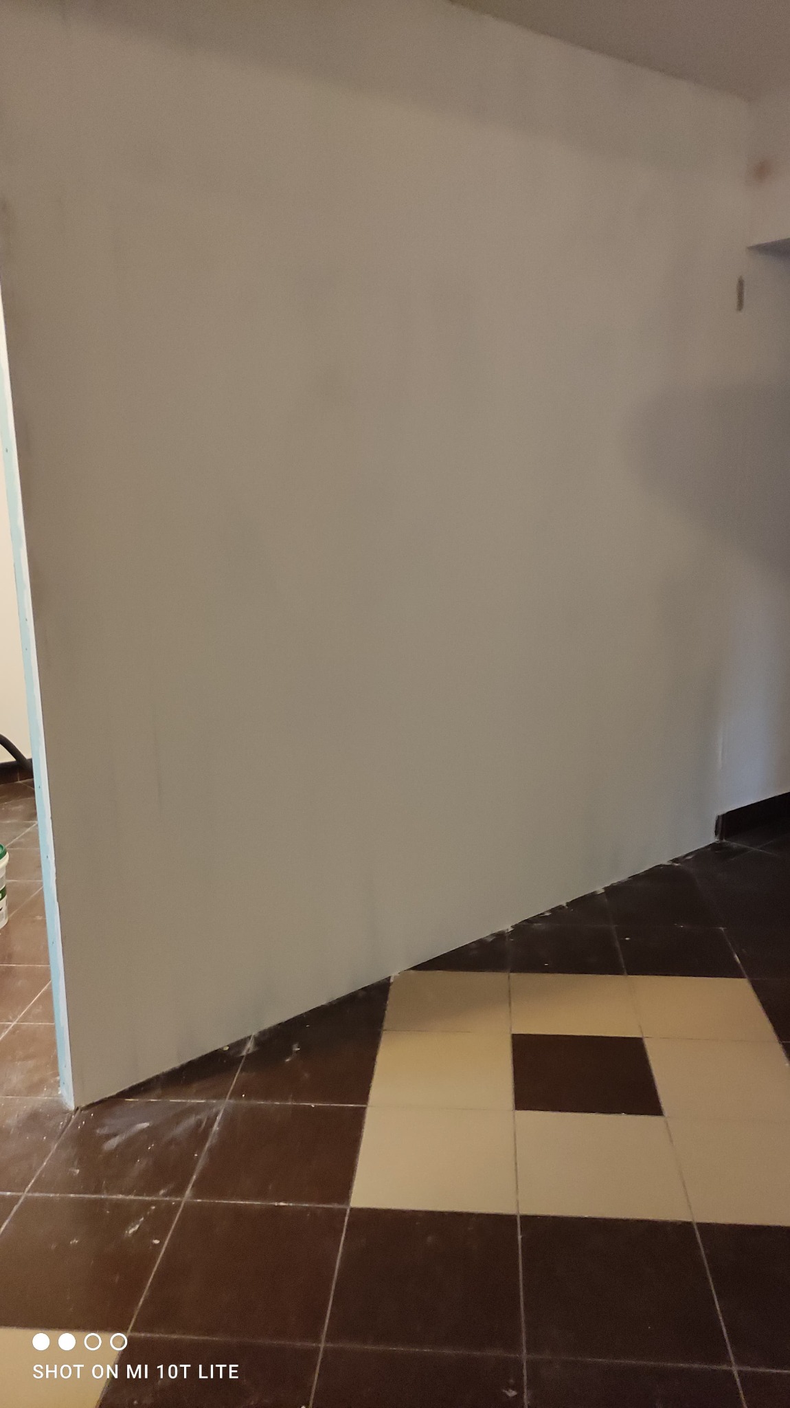 Świeżo zagruntowana ściana z płyty karton gips na tle brązowo-beżowej podłogi w szachownicę, widoczne ślady po malowaniu na podłodze.