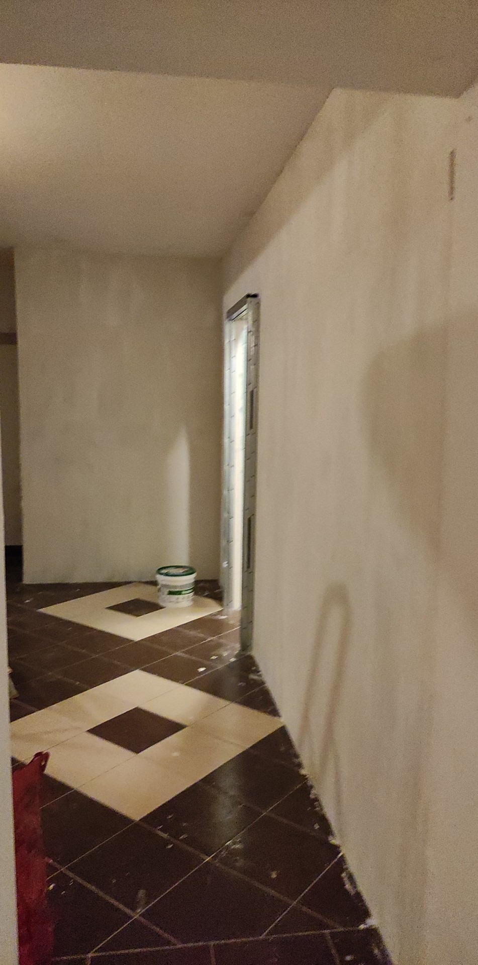 Przygotowane do malowania ściany z widoczną puszką farby w pomieszczeniu z ułożonymi płytkami podłogowymi w karo. W tle ościeżnica drzwiowa bez drzwi.