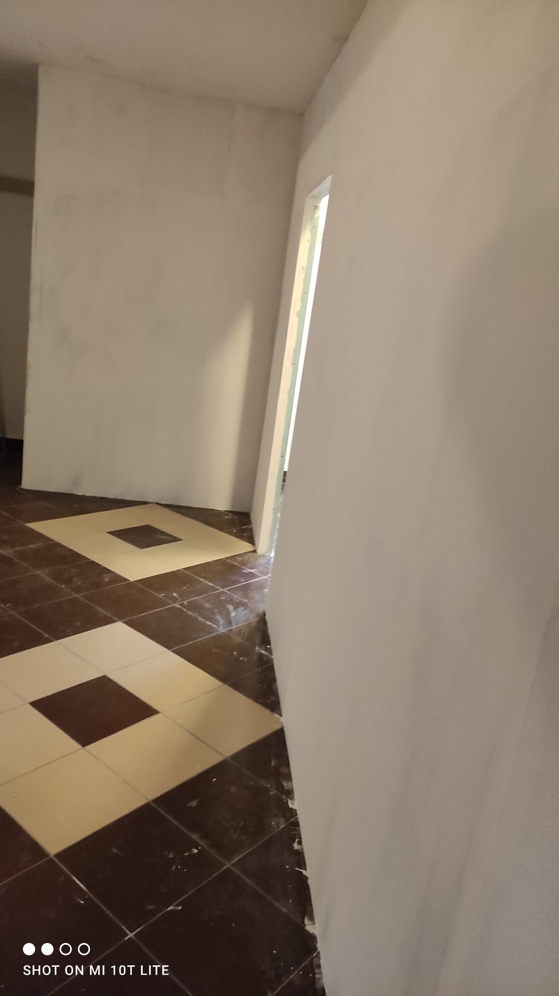 Korytarz w trakcie remontu: świeżo zagruntowane ściany na biało, widoczna stara podłoga z brązowo-beżowych płytek ułożonych w kwadraty, przygotowanie do dalszych prac wykończeniowych.