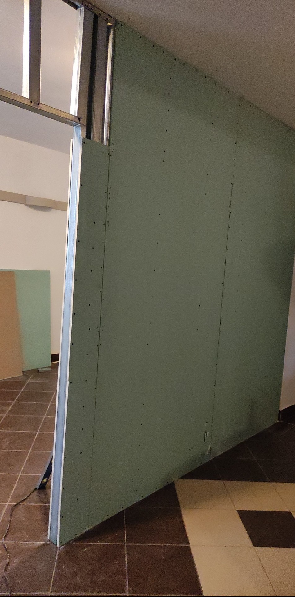 Montaż ściany działowej z płyty kartonowo-gipsowej na metalowym stelażu, widoczne łączenia płyt, szpachlowane otwory po wkrętach, podłoga wyłożona brązowo-białymi płytkami.