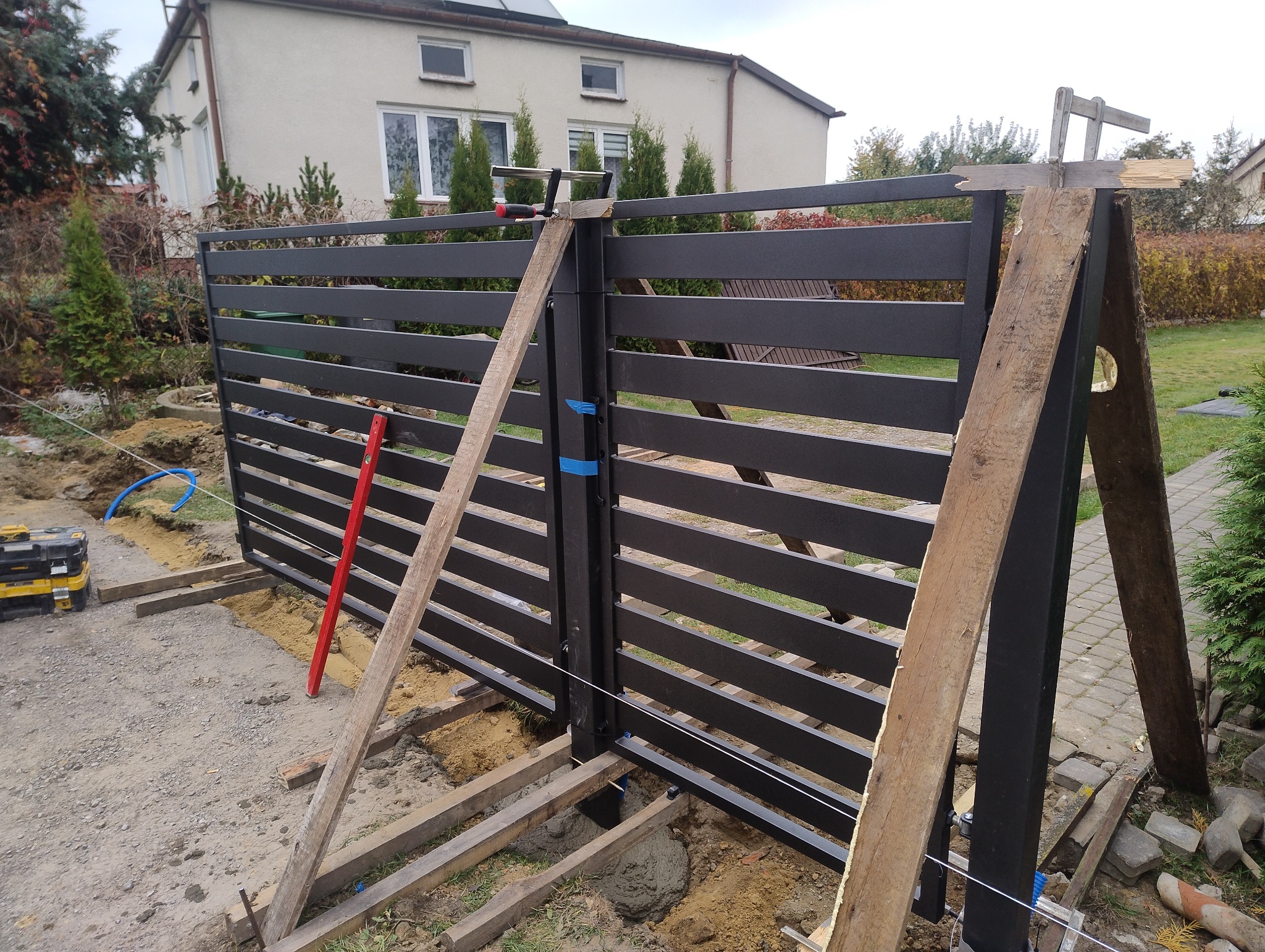 Montaż nowoczesnego ogrodzenia panelowego z poziomych listew, wspartego drewnianymi stemplami. Widoczne narzędzia i elementy konstrukcyjne w trakcie instalacji.