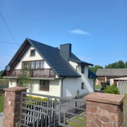 Dom jednorodzinny z ciemnoszarą blachą trapezową na dachu i kominie, balkon z drewnianą balustradą, ogrodzenie z kamiennymi słupkami i metalowymi przęsłami.