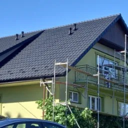 Dom jednorodzinny z nowym, ciemnoszarym dachem z dachówki ceramicznej, widoczne rusztowania przy ścianie szczytowej, sugerujące prace remontowe elewacji.