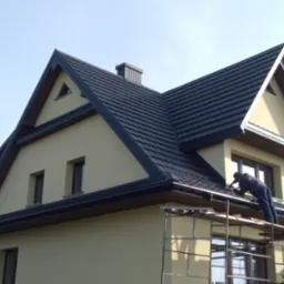 Pracownik na rusztowaniu montuje rynny na nowym, ciemnoszarym dachu domu o jasnej elewacji, widoczne okna i komin.