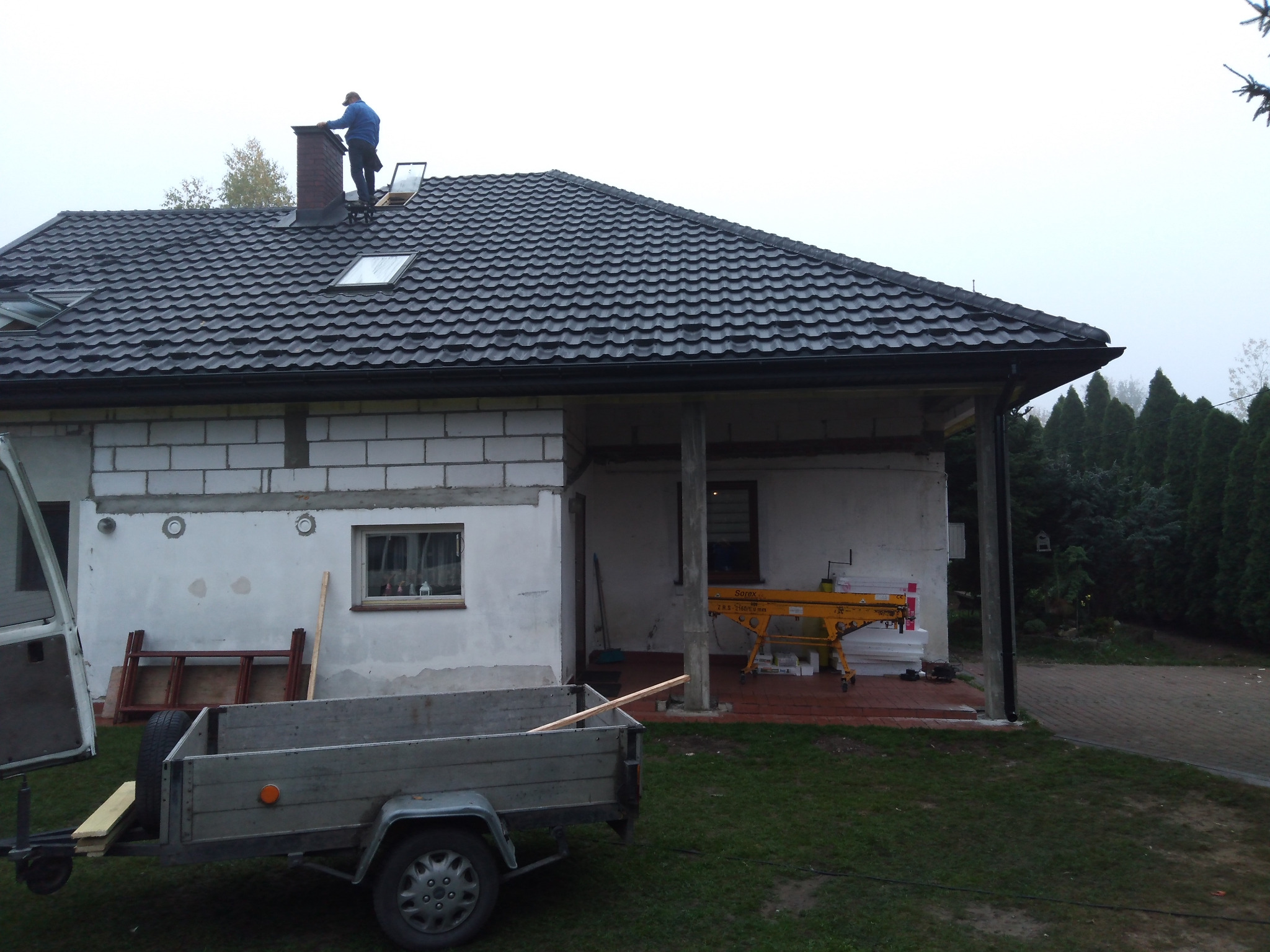 Pracownik w niebieskiej kurtce na dachu domu z ciemną dachówką, widoczne okna dachowe i komin, przyczepka i rusztowanie na posesji.