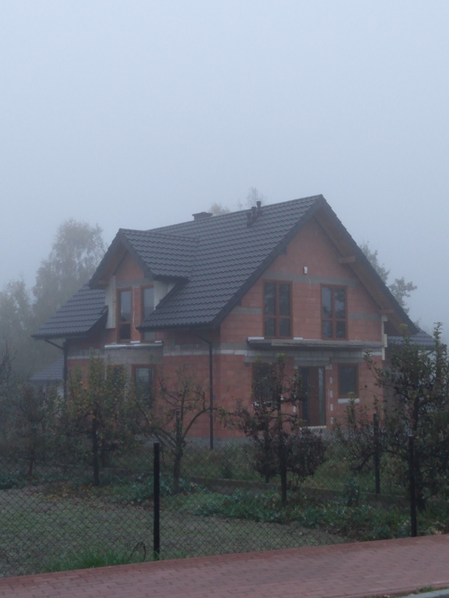 Surowy budynek jednorodzinny z ciemną dachówką w porannej mgle, widoczne niezakończone prace murarskie i stolarka okienna.