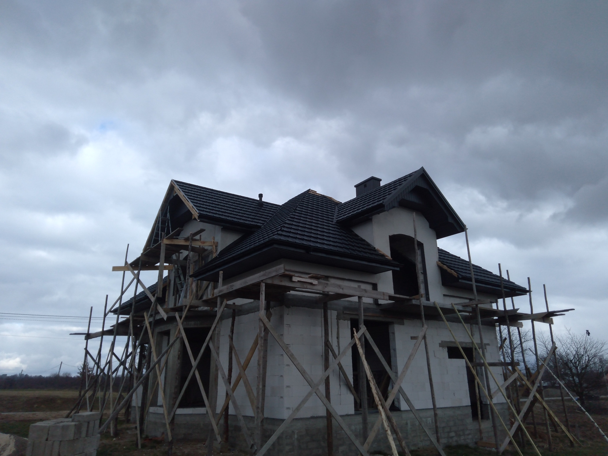 Surowy budynek w trakcie budowy z czarnym dachem, otoczony drewnianym rusztowaniem na tle pochmurnego nieba.