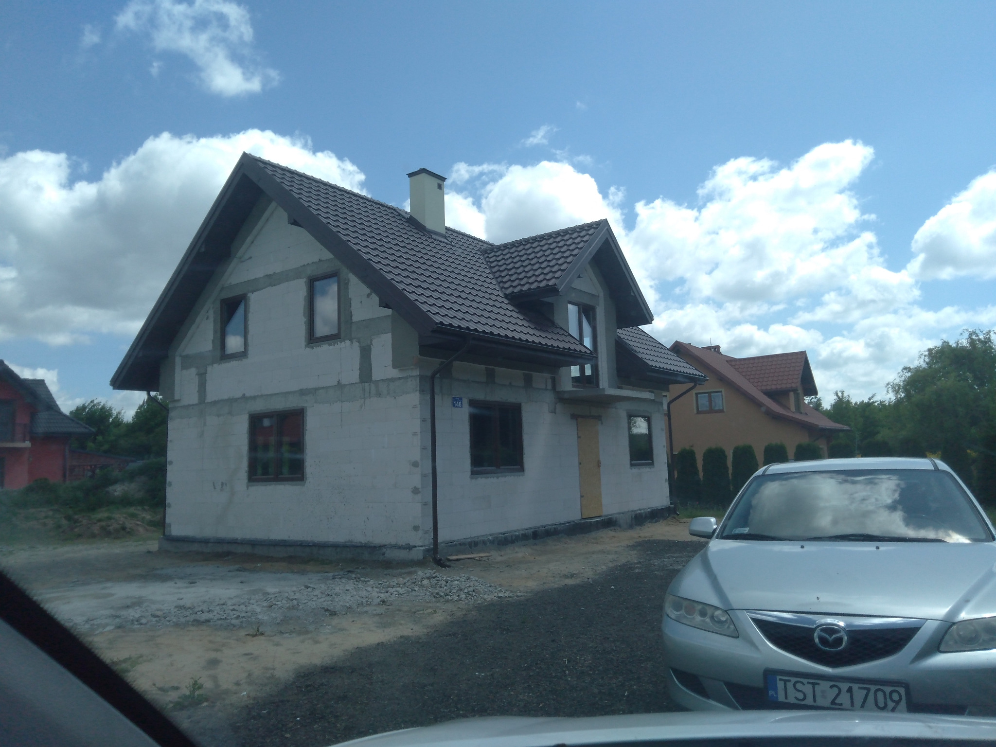 Surowy dom w trakcie budowy z ciemną dachówką, widok zza samochodu, błękitne niebo z chmurami w tle.
