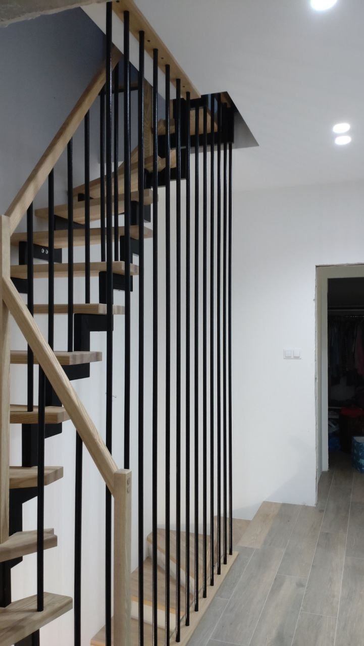 Nowoczesne schody z drewnianymi stopniami i balustradą z czarnych, pionowych prętów, jasne drewno poręczy kontrastuje z czernią elementów konstrukcyjnych, widok z dołu na tle białej ściany i drzwi.