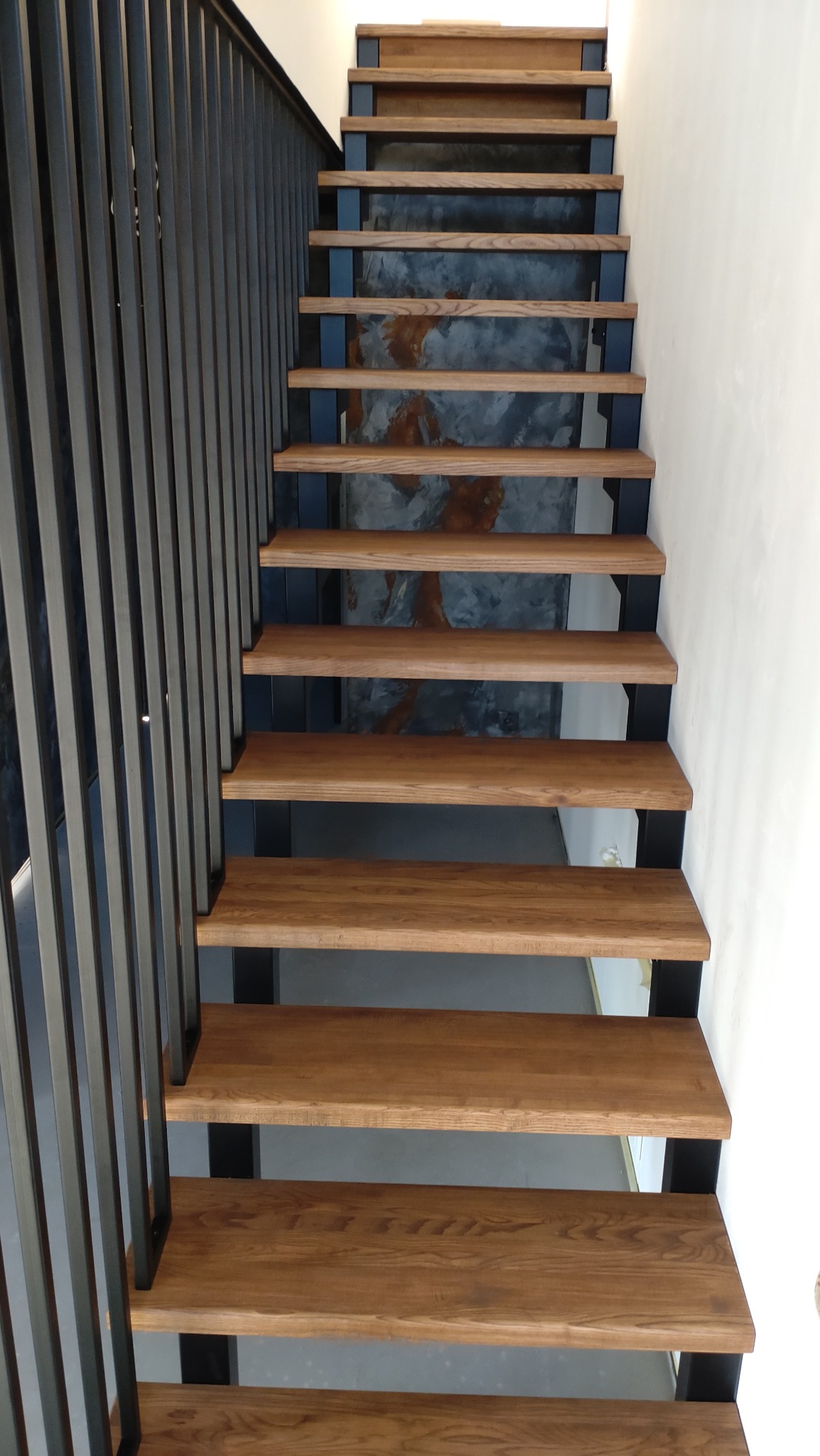 Drewniane schody z metalowymi wspornikami i poręczą, widok z góry, z tłem w odcieniach szarości i brązu imitującym beton architektoniczny.