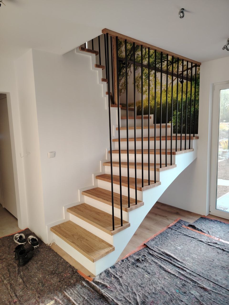 Jasne, drewniane stopnie schodów z białymi podstopnicami i czarną, metalową balustradą z drewnianym poręczą, zabezpieczone folią malarską na podłodze.