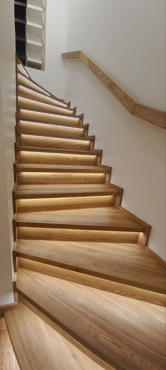 Jasne, drewniane schody z podświetleniem LED pod każdym stopniem, widok od dołu, z fragmentem drewnianej poręczy.