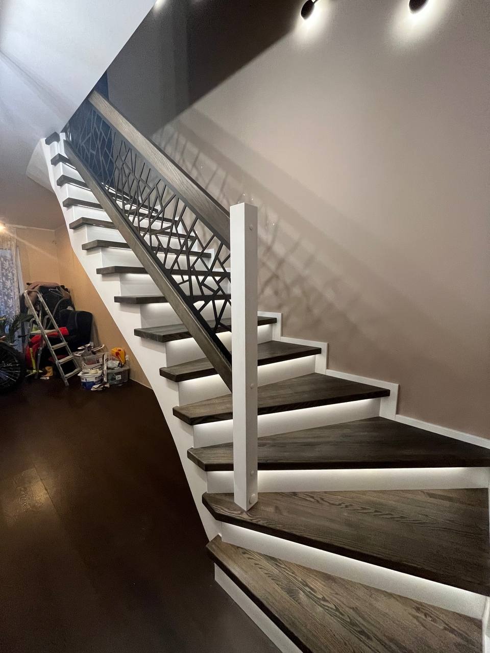 Nowoczesne schody z ciemnego drewna z białymi podstopnicami i podświetleniem LED, ażurowe balustrady z geometrycznym wzorem.