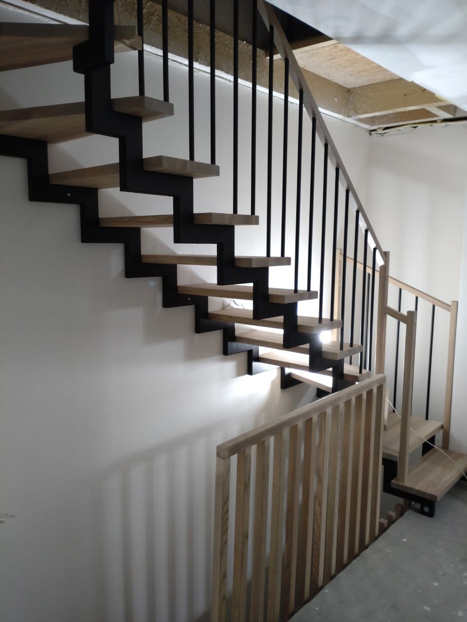 Nowoczesne schody z drewnianymi stopniami i czarną metalową konstrukcją, widoczne niedokończone wykończenie sufitu i fragment drewnianej balustrady.