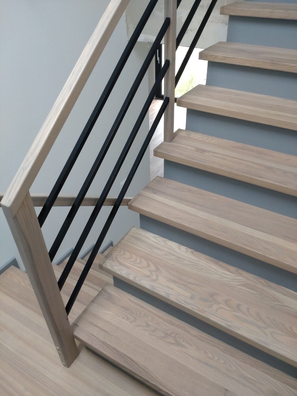 Jasne stopnie z drewna jesionowego z czarną, metalową balustradą o prostych, geometrycznych liniach, osadzone na szarych podstopnicach.